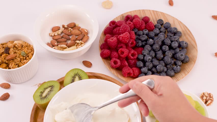 Overhead view of a hand stirring creamy yogurt surrounded by fresh berries, kiwi, and nuts.