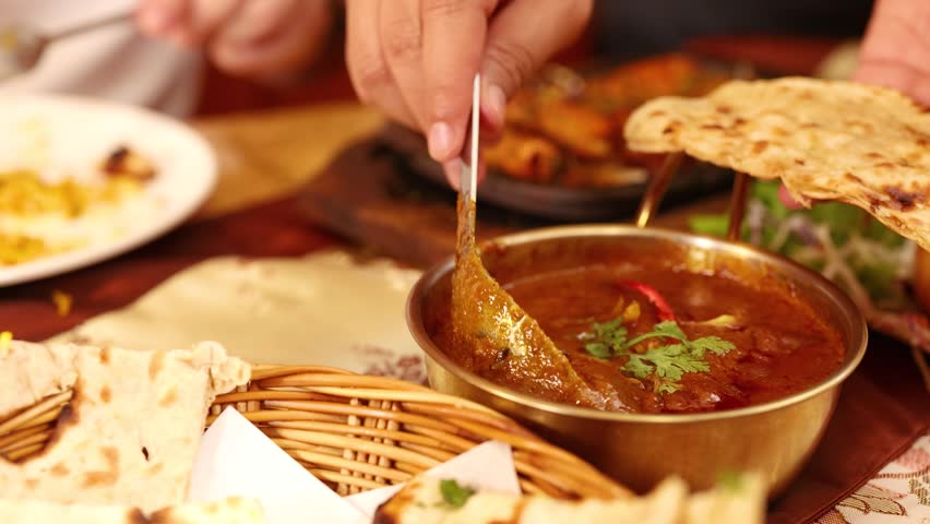 Close-up shot of a hand using a spoon to scoop rich curry from brass bowl.