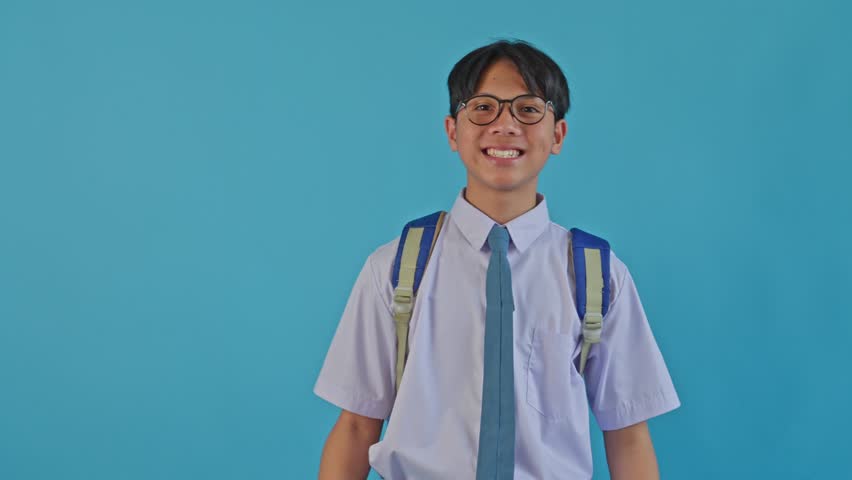 Happy Asian schoolboy in uniform making choosing gesture or pointing finger gun at camera over blue backdrop
