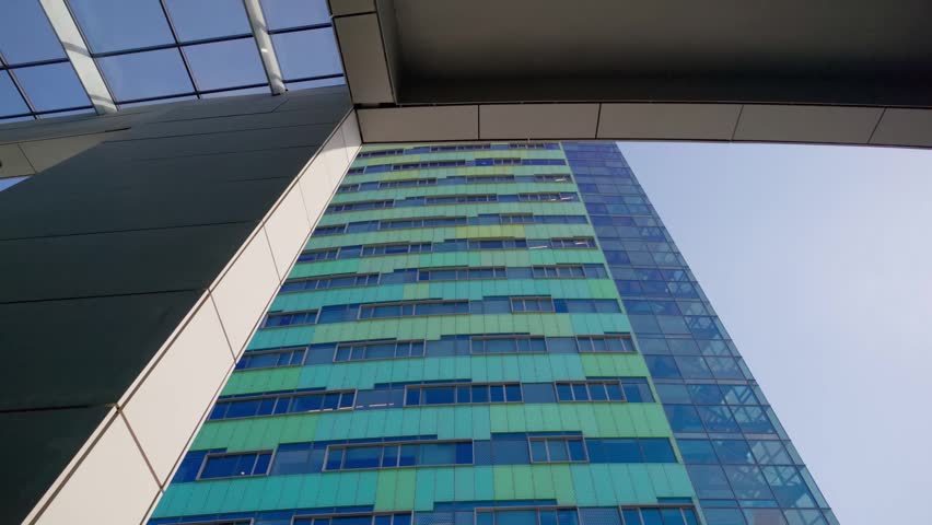 Looking up at a tall modern office building in Arnhem, Netherlands, featuring a distinctive green and blue geometric facade. Architectural structure frames the clear blue sky, emphasizing urban des...