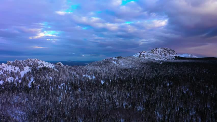 Aerial view of a snowy alpine wilderness with conifer forest rolling hills rocky peaks and dramatic clouds at dusk.