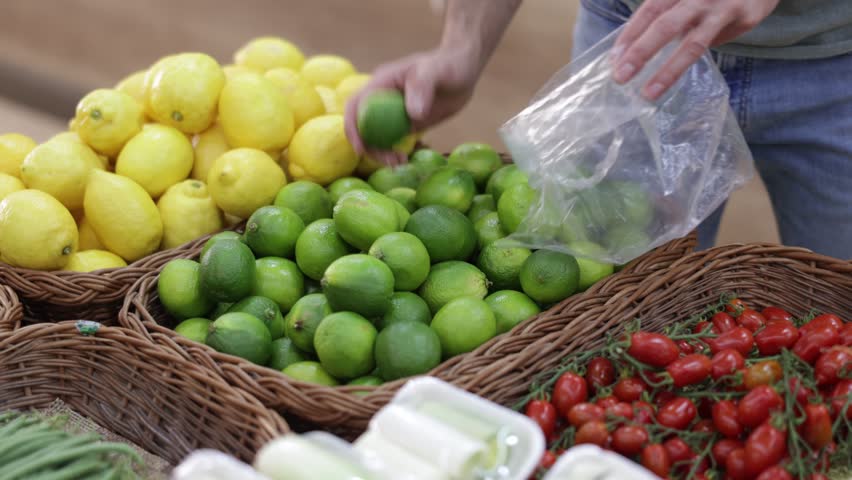 Close up of a shopper hands putting fresh limes in a bag to weigh before buying. Male hands holding lime near fruit baskets. High quality 4k footage
