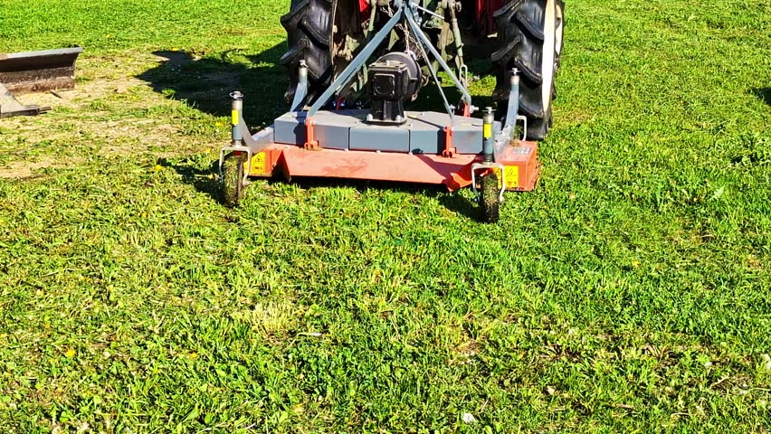 Red agricultural tractor mowing overgrown grass in a large garden during a sunny day