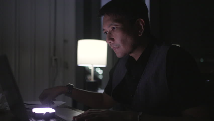Businessman smiles while working late at night at home, using a laptop and talking on a mobile phone, illuminated by a table lamp in a cozy apartment.