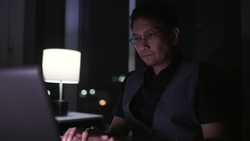 Businessman smiles while working late at night at home, using a laptop and talking on a mobile phone, illuminated by a table lamp in a cozy apartment.