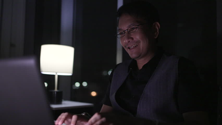 Businessman smiles while working late at night at home, using a laptop and talking on a mobile phone, illuminated by a table lamp in a cozy apartment.