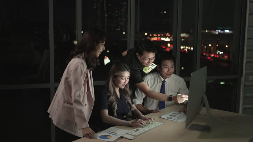 Multiethnic business team collaborates late at night in a modern office, discussing charts and data on a computer screen with focus, communication and teamwork.