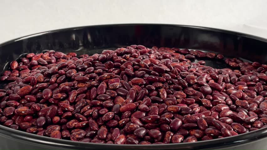 Slow motion close up of human hands scooping raw red kidney beans from a black tray, raw speckled adzuki beans falling through fingers, healthy organic food preparation concept.