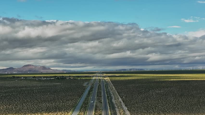 Aerial view shows long roads and open land in the USA. Fields stretch out on both sides with mountains on the horizon. Clouds gather in the sky above the landscape.