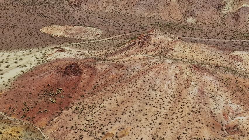 This aerial shot shows a wide view of a desert landscape in the USA. Brown hills create patterns on the ground while scattered small plants dot the surface under bright sunlight.