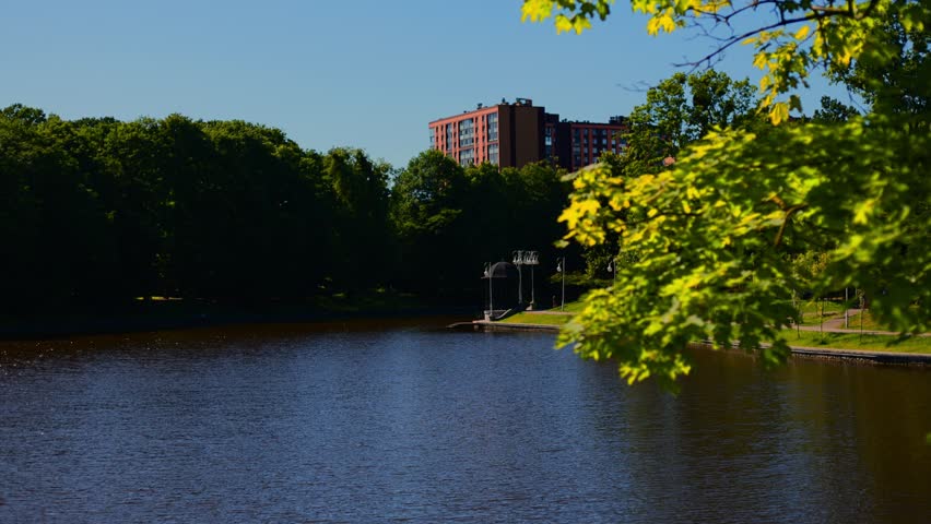 Scenic lake view surrounded by lush green trees and urban architecture. Summer park landscape with clear blue sky. Nature recreation area for outdoor relaxation.