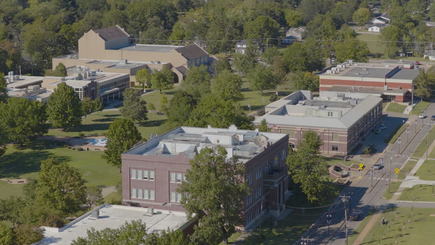 College campus buildings surrounded by lush green trees and lawns under clear midday light in Clarksville, Arkansas, with a quiet street visible.