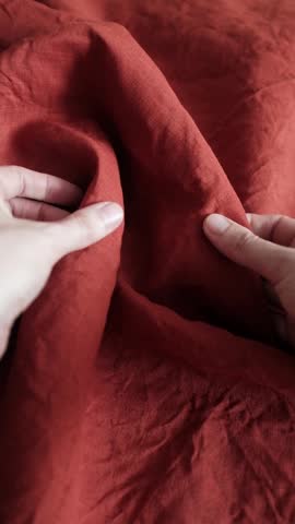 Female hands gently feeling and assessing the premium quality of a crumpled terracotta red linen fabric. A woman runs her fingers along the natural weave and organic texture of the rustic, pure flax textile, showcasing its soft drapes and wrinkled aesthetic. Perfect for themes of home decor, tailoring, sustainable fashion, clothing manufacturing, and high-quality organic materials.