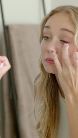 Vertical video: Gazing into mirror, woman in tank doing eye care at home using fingers for symmetry. Skincare, grooming, reflection, bathroom, vanity, softlit, vertical video