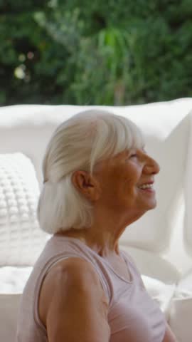 Vertical video: Waving senior woman replying to offscreen wave, sitting on sofa with plants in tank. Elderly, couch, sunroom, daylight, greenery, cheerful, vertical video