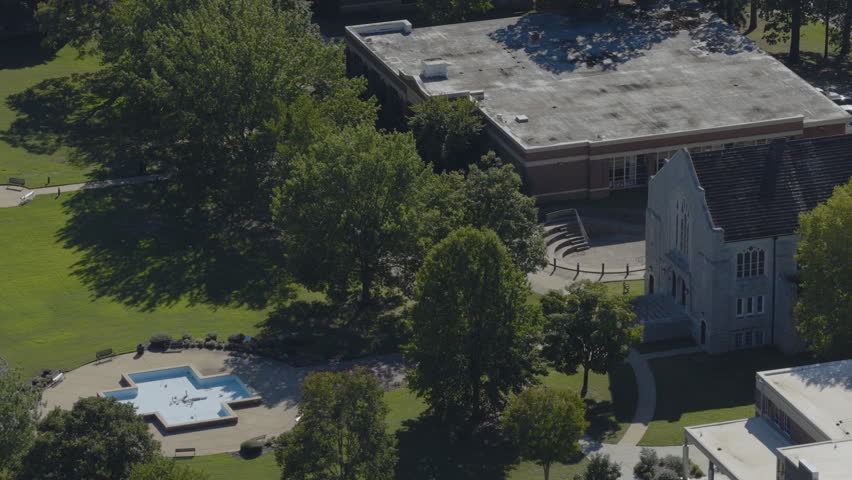 Aerial drone footage captures a college campus in Clarksville, Arkansas, featuring a central fountain, green trees, and academic buildings under clear midday light.