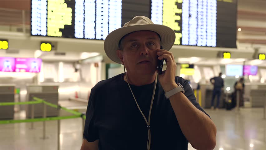 Senior traveler in a hat having a phone conversation while waiting for his flight at the airport