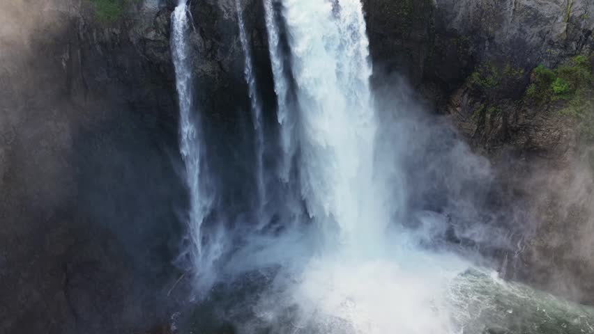 Snoqualmie Falls plunges through misty basalt cliffs as historic lodge perches above churning whitewater amid dense evergreen rainforest canopy below. Washington, USA. Aerial drone rise shot
