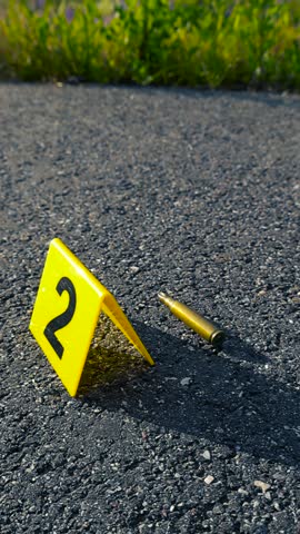 Yellow crime scene evidence marker with the number two next to a spent rifle bullet casing lying on the asphalt road, representing forensic investigation at the site of a shooting incident