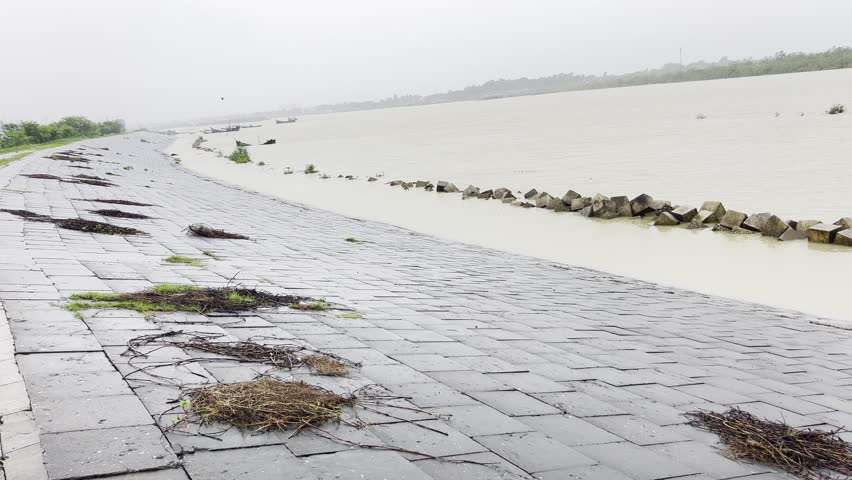 A calm river overflow scene with rising water spreading across the ground near a stone walkway under a cloudy sky.