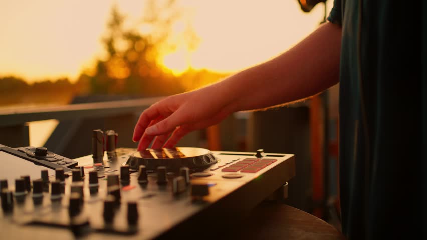 energetic dj adjusting sound mixer controls at sunset over wooden bridge with trees in the background. vibrant outdoor performance setting with golden yellow and orange tones.