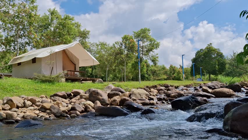 A scenic view of a luxury glamping tent pitched beside a clear, fast-flowing river with a rocky bed, surrounded by lush green trees and a bright blue sky on a sunny day.