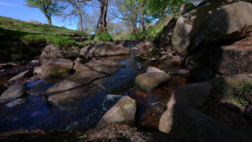 A static view of Burbage brook flowing through Padley Gorge surrounded by rocks, and parkland in the background in Derbyshire, UK