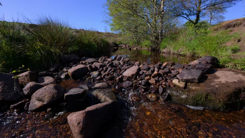 A static view of Burbage brook flowing through Padley Gorge surrounded by rocks, and parkland with blue sky in the background in Derbyshire, UK