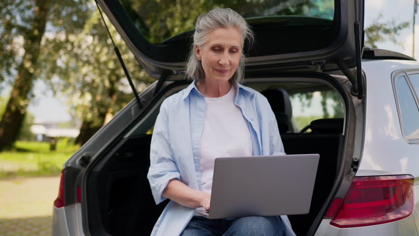 Cheerful happy Caucasian old aged business woman working laptop sitting in car trunk smiling carefree senior mature retired female using computer distant near auto outdoors park nature near automobile