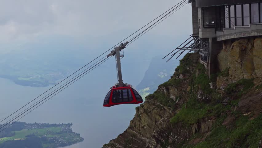 Red cable car descends from Mount Pilatus above Lake Lucerne in Switzerland, with rocky cliffs, misty Alps, and clouds creating a dramatic mountain travel scene. 4K UHD video.