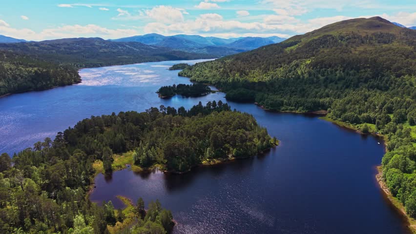 Aerial drone view over Loch Beinn Mheadhoin in Glen Affric, Scotland, with forested islands, blue water and Highland mountains under a bright sky. 4K UHD video.