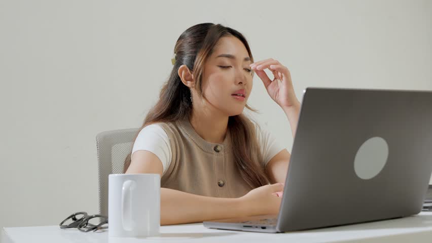 Confused young Asian female employee is working using her laptop while feeling headache and stressful.
