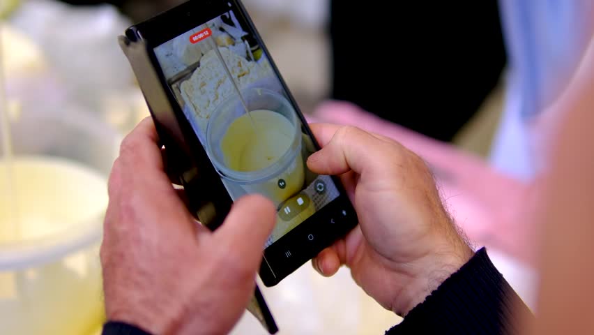Male hands filming fresh farm cream in metal bucket with smartphone while market vendor scoops homemade dairy product at traditional farmers market