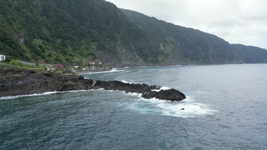 Aerial 4K drone view of rugged green mountain cliffs and a small village along a wild volcanic coast with waves crashing on black rocks on the Atlantic Ocean in Madeira, Portugal. Moody north coast.
