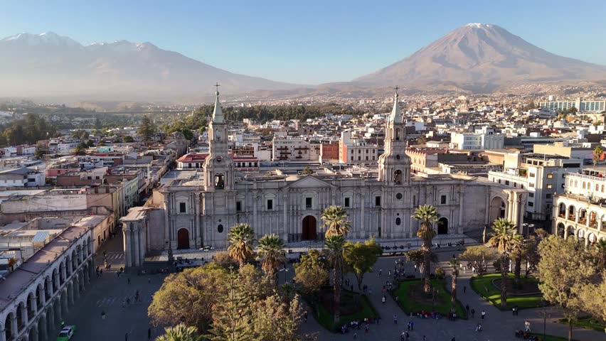 An ascending aerial shot reveals the historic Basilica Cathedral of Arequipa in Peru, with the majestic El Misti volcano dominating the background.