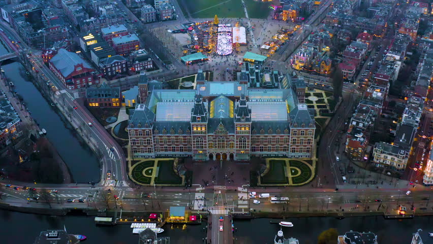 Aerial night view of the Rijksmuseum in Amsterdam, Netherlands, with an ice skating rink, Christmas tree and holiday market on Museumplein, canal boats and city traffic at dusk in winter.