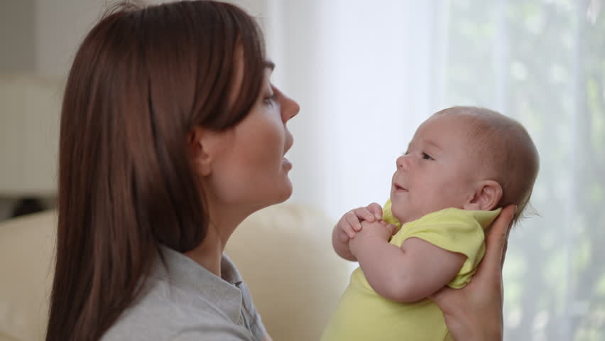 Loving young mother holding her newborn baby in her arms. Woman gently kissing and hugging her cute infant son, expressing tenderness and care