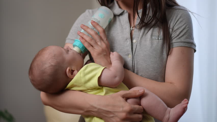Caring mother holding her adorable infant child in her arms while feeding with formula from a bottle. Concept of motherhood and childcare