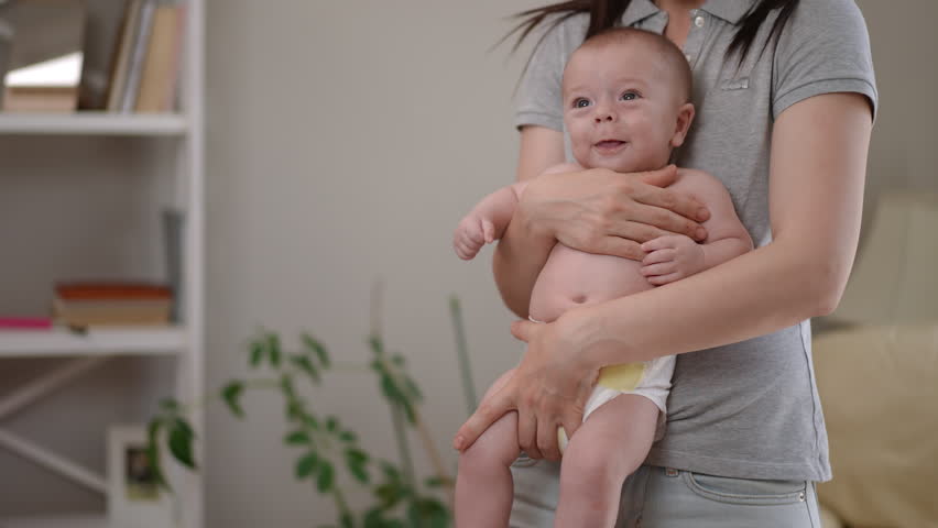 Loving mother tenderly holding her cute newborn baby boy in diapers. The infant is smiling, looking around, and expressing emotions