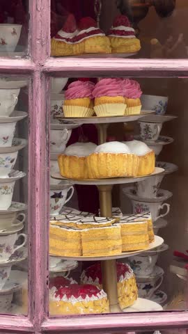 Assorted cakes and muffins displayed on a tiered stand in a shop window. Vintage floral teacups stacked beside delicious pastries. Bakery showcase for dessert cafe business concept.