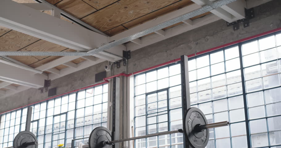 Camera is tilting down, revealing weight bench and loaded barbell on squat rack, showing loft gym. Industrial, daylight, windows, plants, chairs, mats, fitness