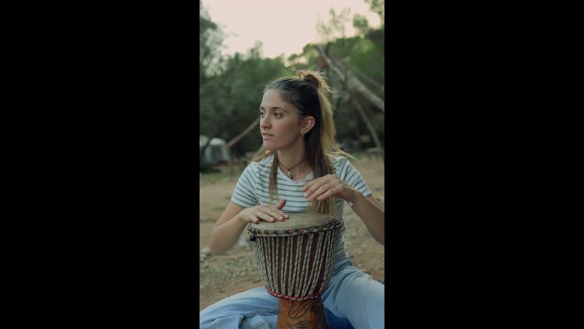 Young woman playing a traditional djembe drum while sitting outdoors in a natural setting