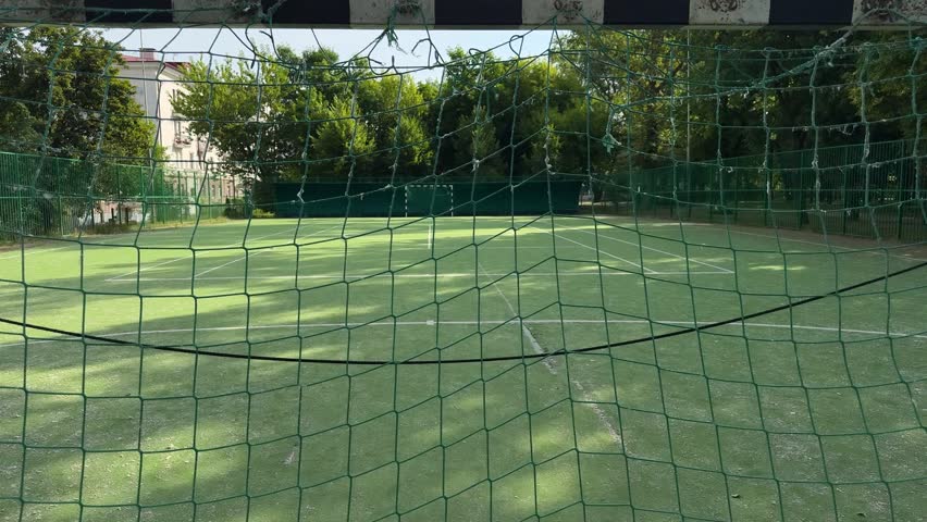 View through green net at empty artificial turf football court. Outdoor mini soccer field with goal, trees, summer sports facility.