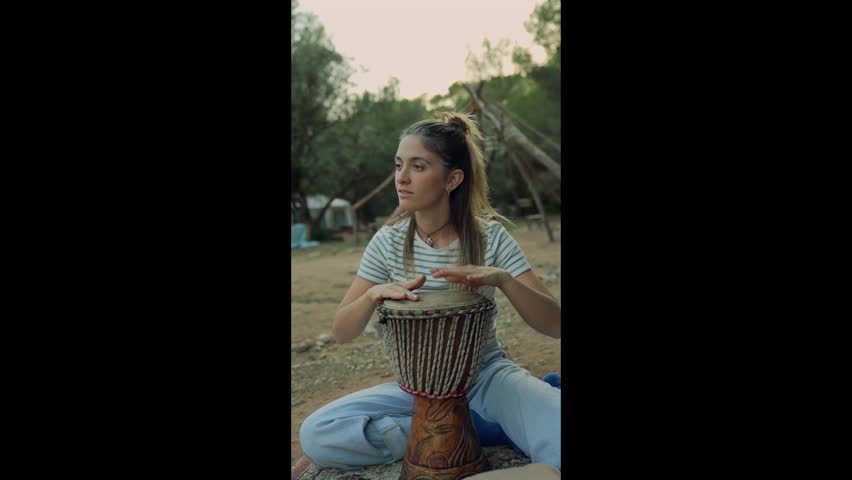 Young woman playing a traditional african djembe hand drum while sitting outdoors in nature