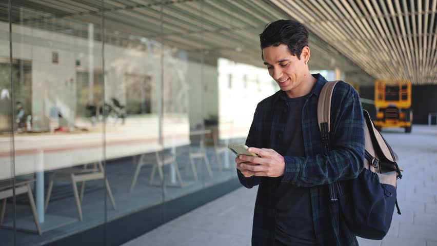 Young hispanic man walking while using mobile phone. Happy university student in the city