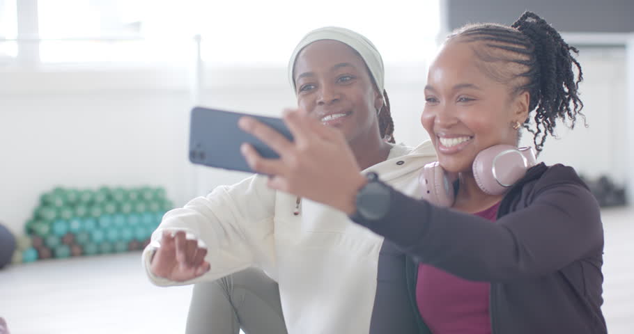 2 African women friends in workout clothes posing for selfie with phone in studio scrolling selfies. Fitness, gym, duo, backlit, laughter, headphones, braids
