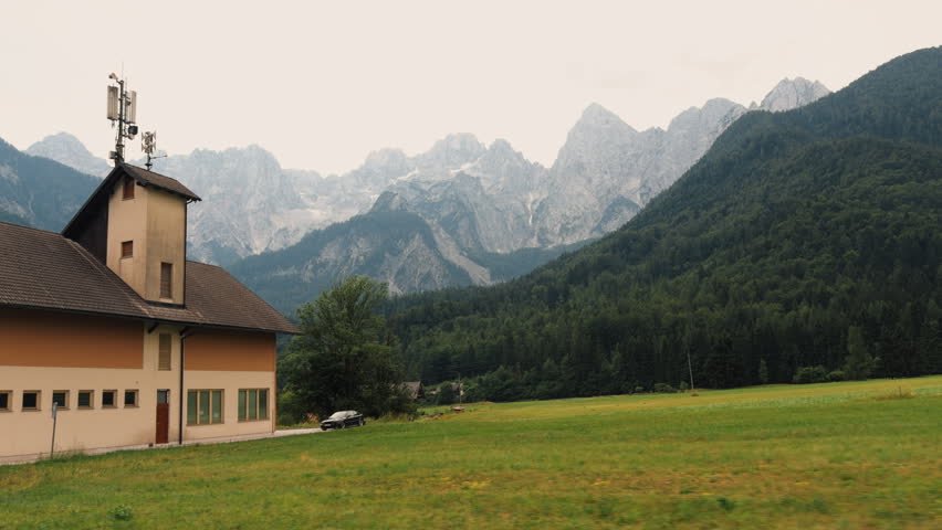 A rural fire station building stands beside a grassy field with alpine mountains and forest in the background. Antennas rise from the roof under a pale cloudy sky.