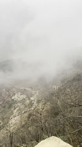 Fog and low clouds rolling over rocky mountain valley. Misty landscape with bare trees and terraced hills in background. Concept of nature, mountains, fog, wilderness, dramatic weather, and rural terrain