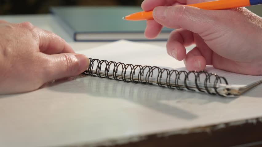 Hand writes with ballpoint pen in spiral-bound notebook on sheet of paper while sitting at table close-up