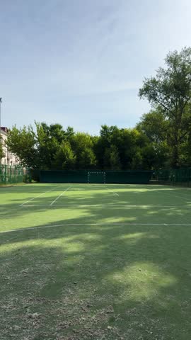 Empty green artificial turf football pitch surrounded by fence and trees under blue sky. Outdoor sports facility, summer day.